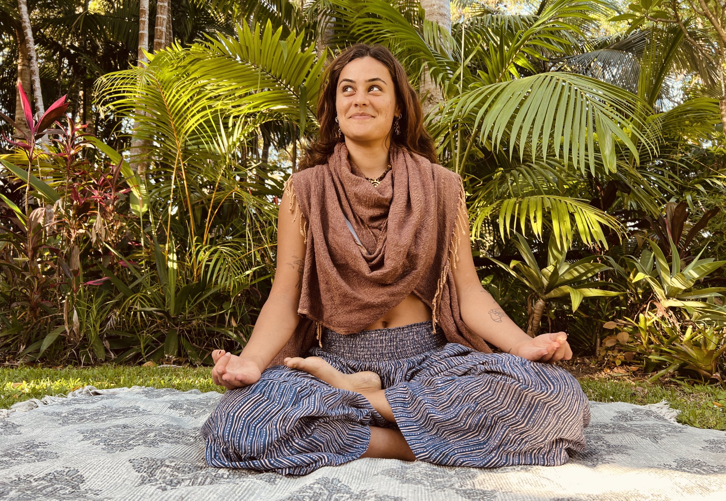 A woman practicing yoga outdoors on a patterned mat in a lush tropical garden, sitting cross-legged with a relaxed expression.