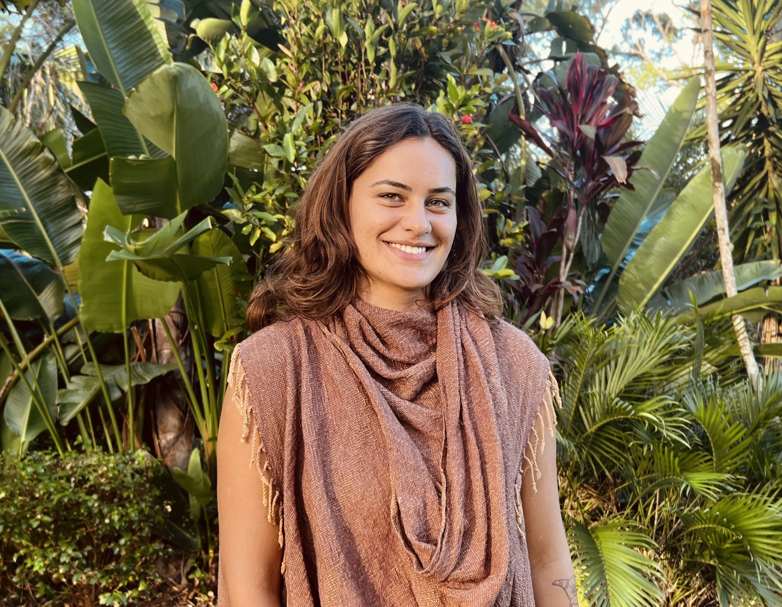 A young woman with wavy brown hair smiling outdoors in a lush green jungle-like setting, wearing a brownish-orange textured sleeveless top and a matching scarf.