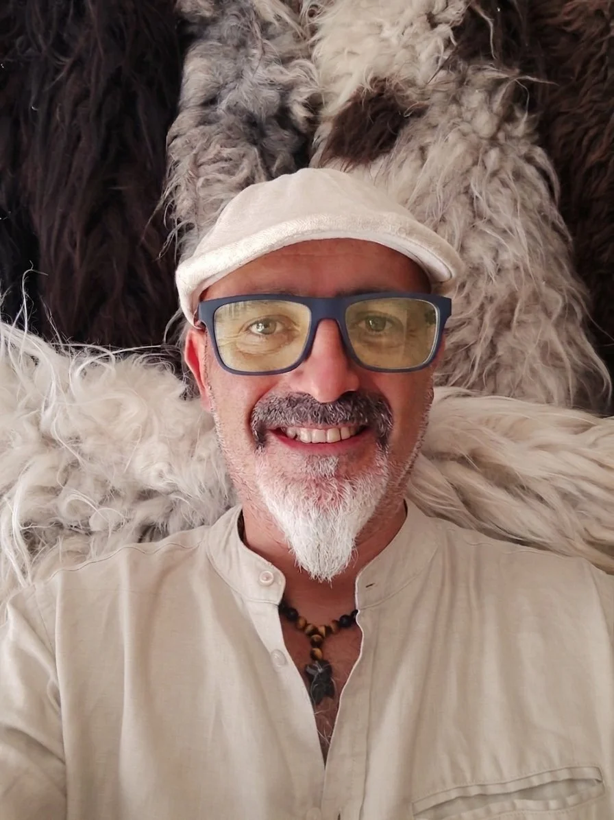 Un uomo sorridente con barba e occhiali, indossa un cappello e una collana, sdraiato su un tappeto di pelliccia.