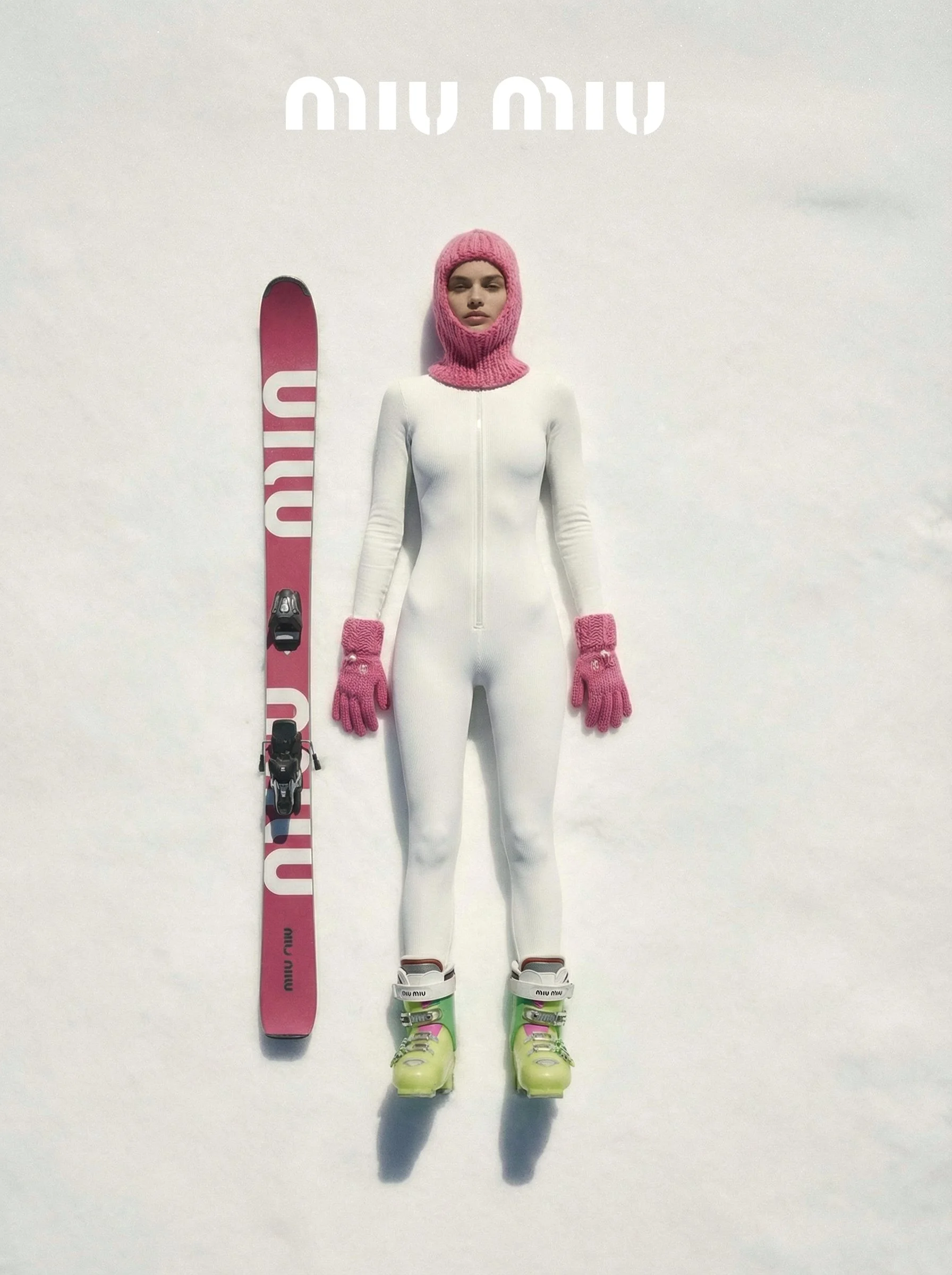 A mannequin dressed in a white ski suit with pink knit accessories, yellow ski boots, a pink knit hat covering the head and neck, pink gloves, and a pink and white Miu Miu ski toboggan positioned vertically against a snow background.