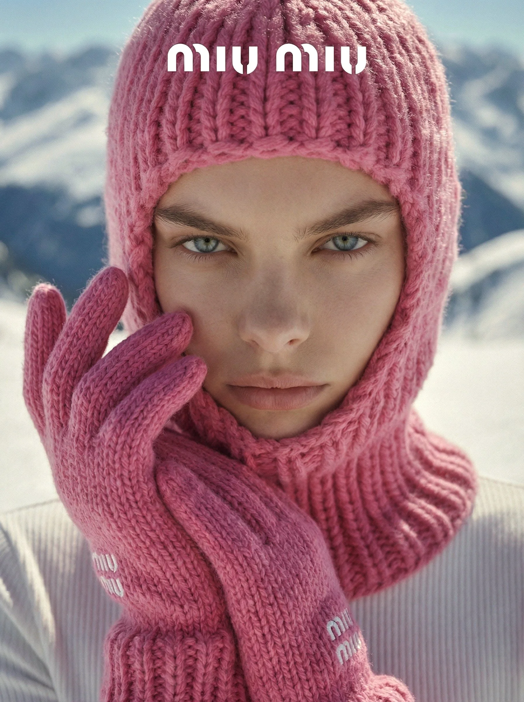 A woman with blue eyes wearing a pink knit winter hat and gloves, surrounded by snow-covered mountains.