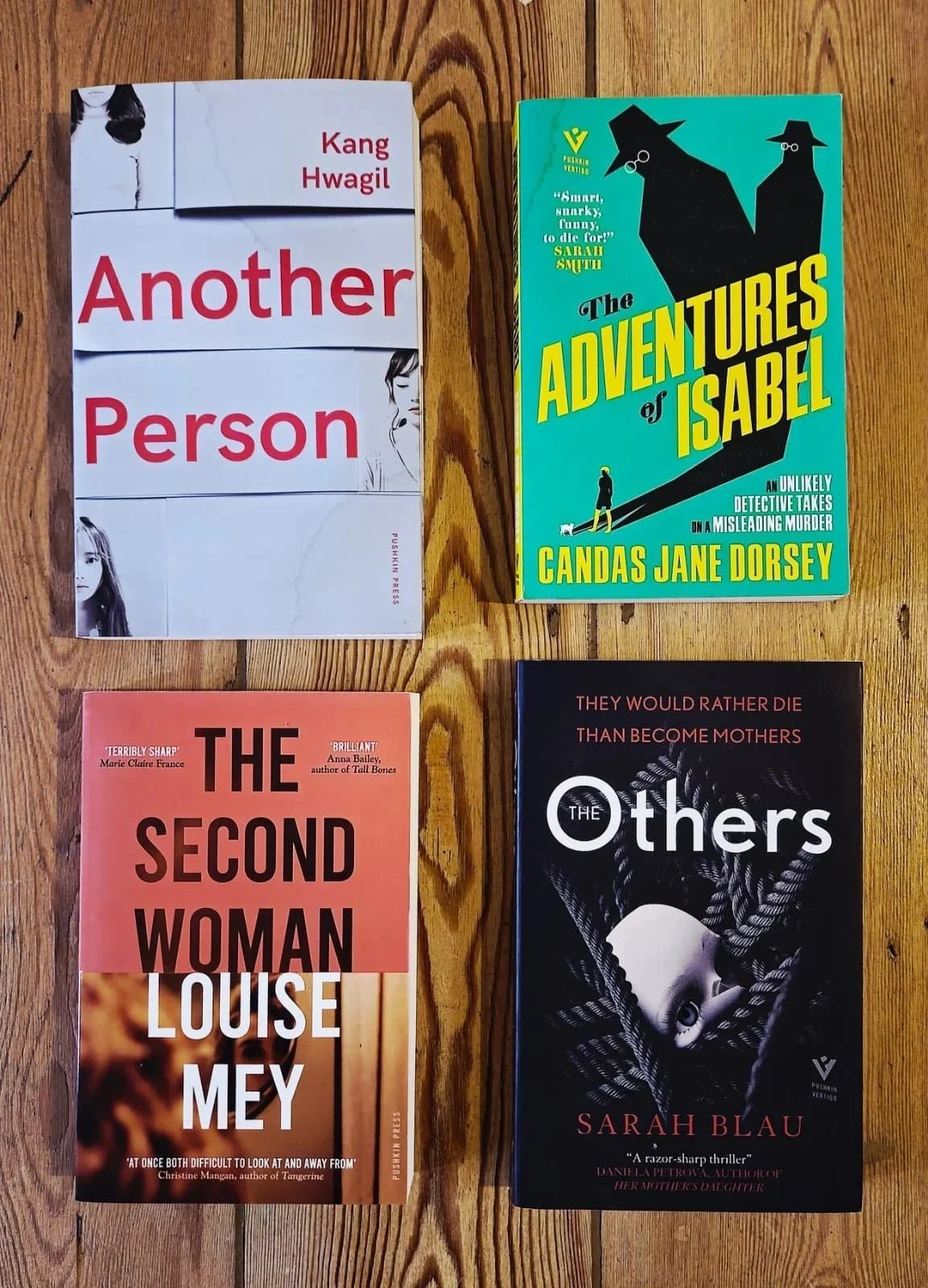 Four books are laid out on a wooden surface. The top left book is titled 'Another Person' by Kang Hwagil, with a white cover featuring black-and-white images of women. The top right book is titled 'The Adventures of Isabel' by Candas Jane Dorsey, with a blue cover featuring black silhouettes of dogs and a small figure walking a dog. The bottom left book is titled 'The Second Woman' by Louise Mey, with a pink-orange cover and a small image of a woman's face. The bottom right book is titled 'The Others' by Sarah Blau, with a black cover featuring a photo of a face partially wrapped in black strings.