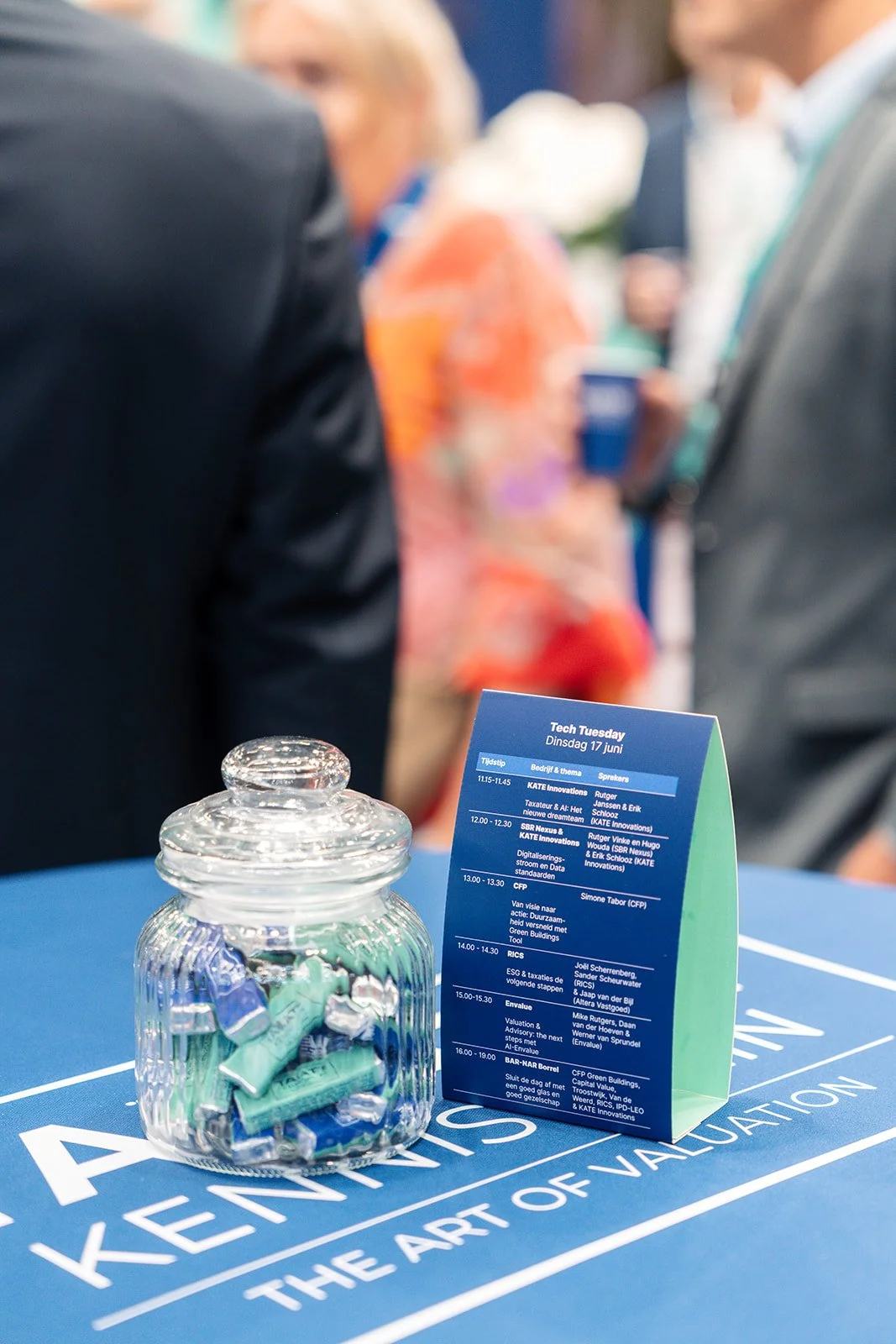 Een blauwe tafel met een glazen doosje gevuld met eurobiljetten en een informatiestandaard met het programma van Tech Tuesday in het Nederlands.