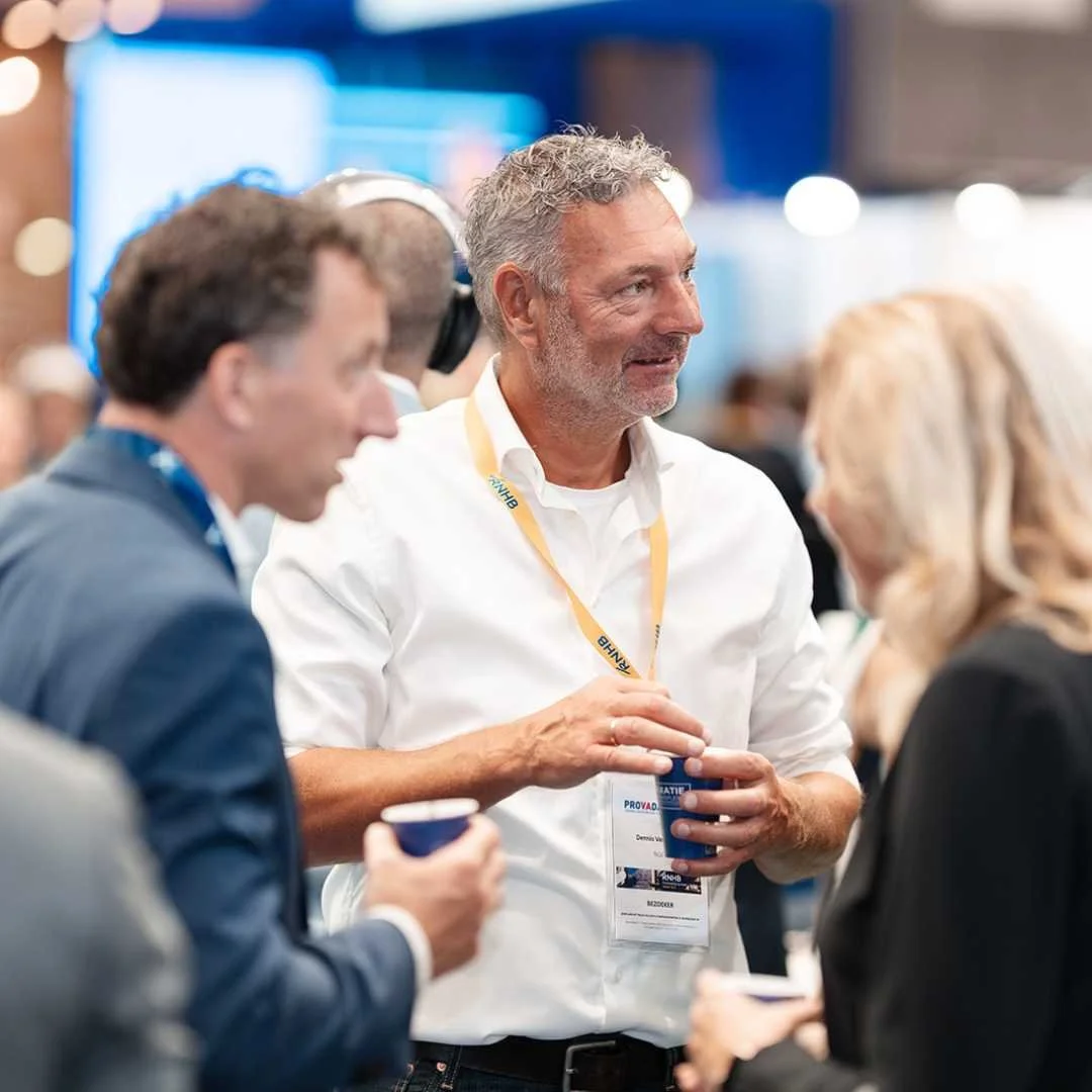 Mensen in gesprek tijdens een evenement, man met grijs haar en witte overhemd, aanwezigen met badges, cafébekers in handen.