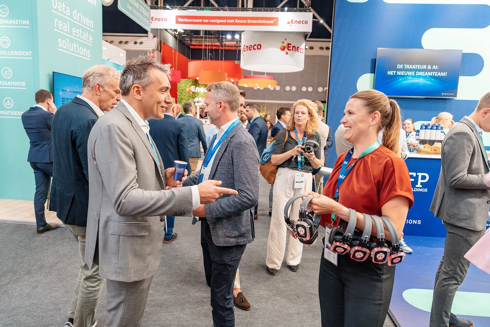 Mensen die praten en netwerken op een beurs met banners en displays, één persoon houdt een set hoofdtelefoons met draagbanden.