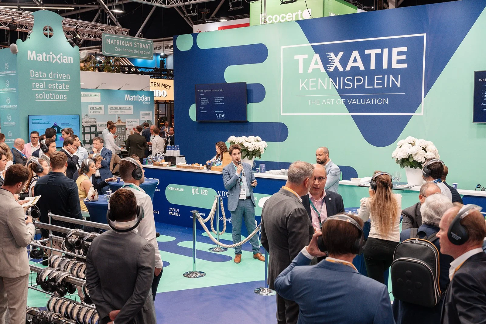 Beursstand met de naam 'TAXATIE KENNISPLEIN' waar een speaker in een grijs pak spreekt voor een publiek.
