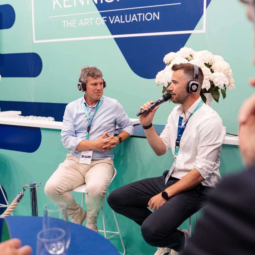Twee mannen met hoofdtelefoons voeren een interview uit op een beurs of conferentie, met een achtergrond die zegt 'The Art of Valuation' en een grote bloemarrangement.