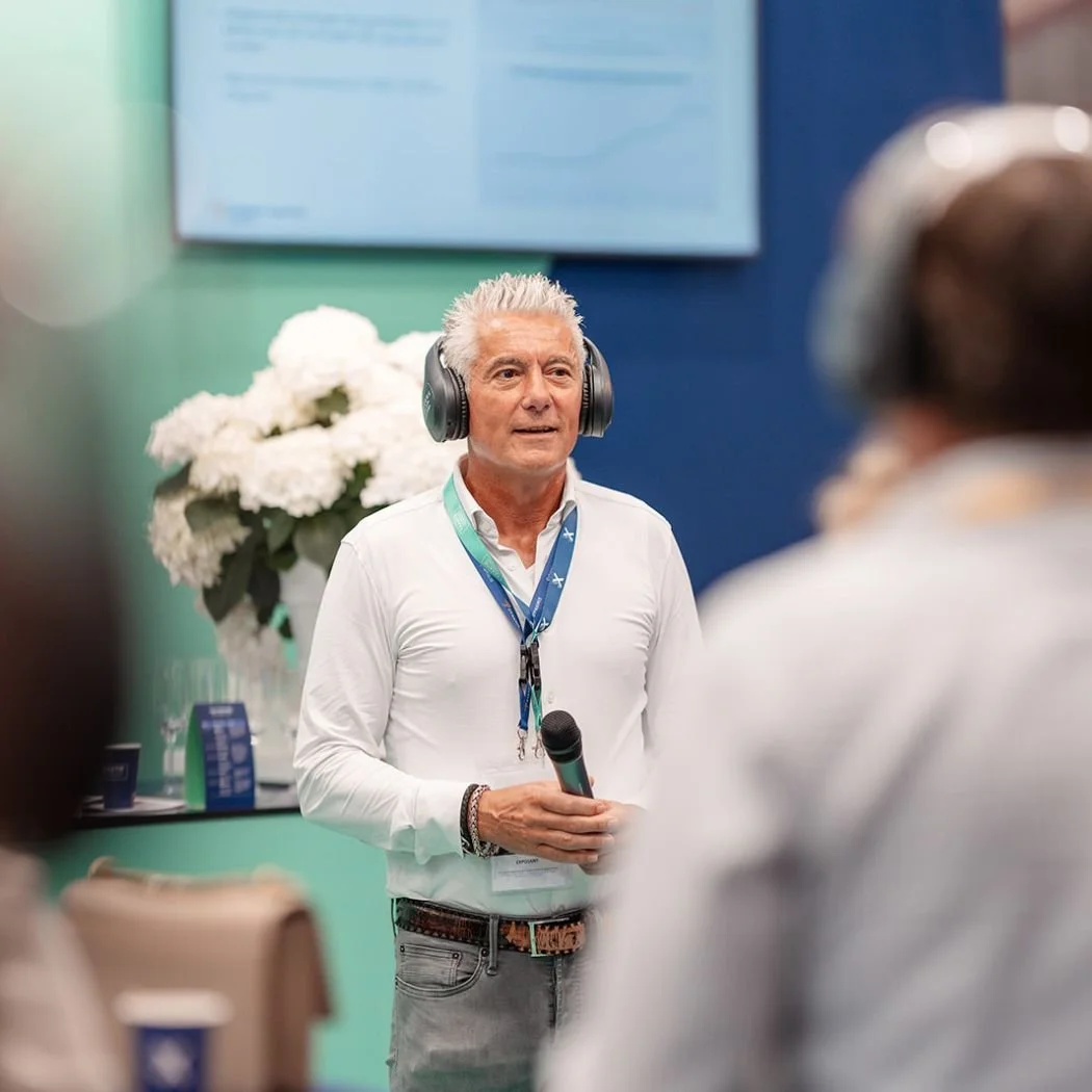 Man met witte haar en witte overhemd houdt microfoon, draagt een headset, staat voor een publiek, achtergrond met bloemen en een scherm met grafieken.