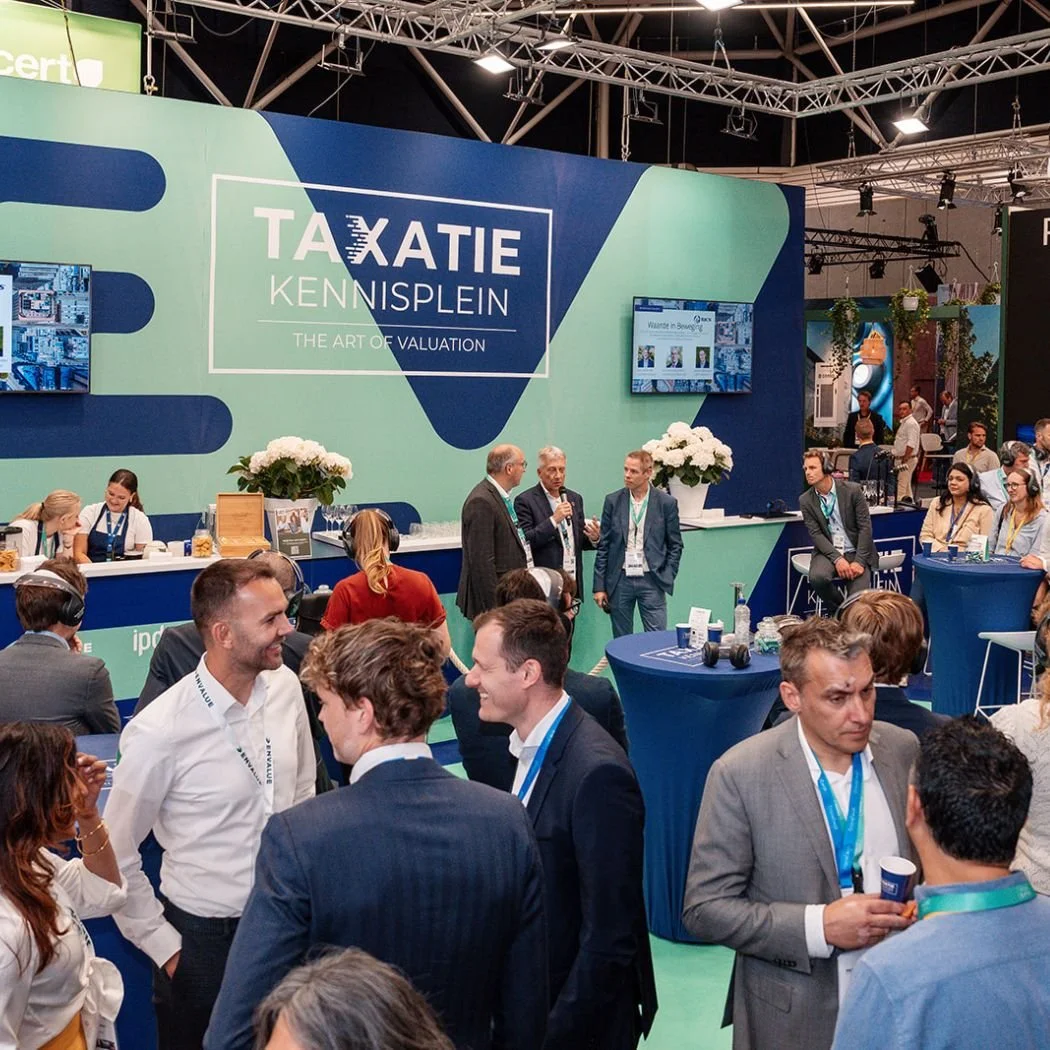 Beurs met een groene en blauwe stand met 'Taxatie Kennisplein' en mensen die praten en netwerken.