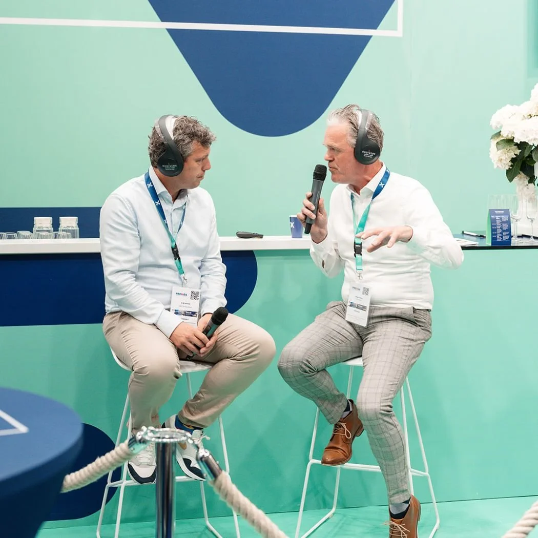 Twee mannen in gesprek met koptelefoons en microfoons, zittend op hoge stoelen, op een groene achtergrond met diagrammen, op een beurs of conferentie.