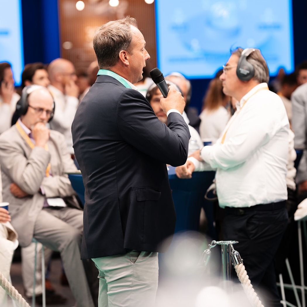 Man in een donker jasje houdt een microfoon vast en praat met een andere man die koptelefoon draagt, omringd door mensen op een congres of conferentie.