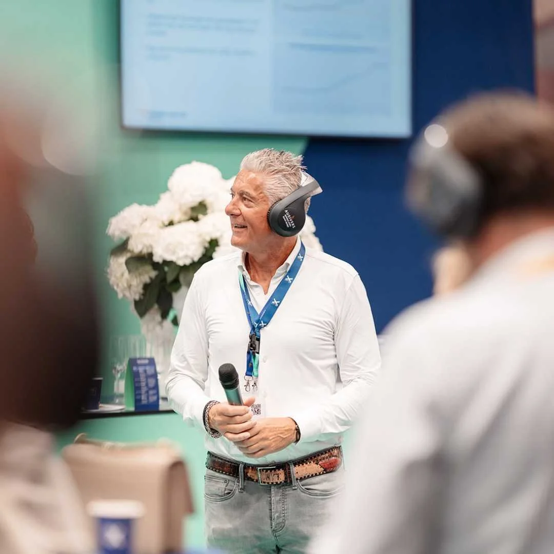Man met grijs haar, een witte blouse, een medaille en een microfoon in een conferentie of presentatie, met een scherm en bloemen op de achtergrond, terwijl hij een presentatie of toespraak houdt.
