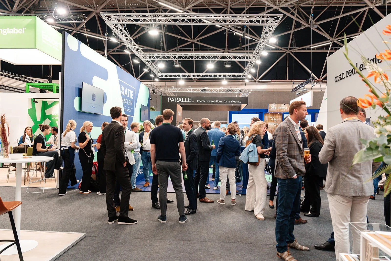 Bezoekers op een beurs of conferentie, met standen en displays, onder een hoog plafond met metalen draagstructuren, mensen die praten en networking.