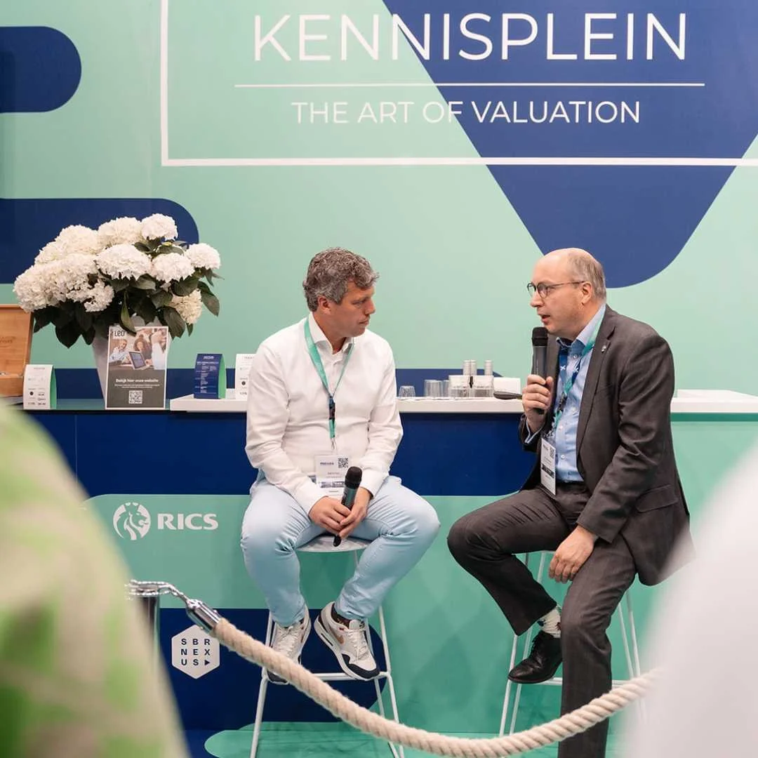 Twee mannen zitten en praten tijdens een paneldiscussie op een beurs, op de achtergrond een groene muur met de tekst 'KENNISPLEIN THE ART OF VALUATION' en het RICS-logo.