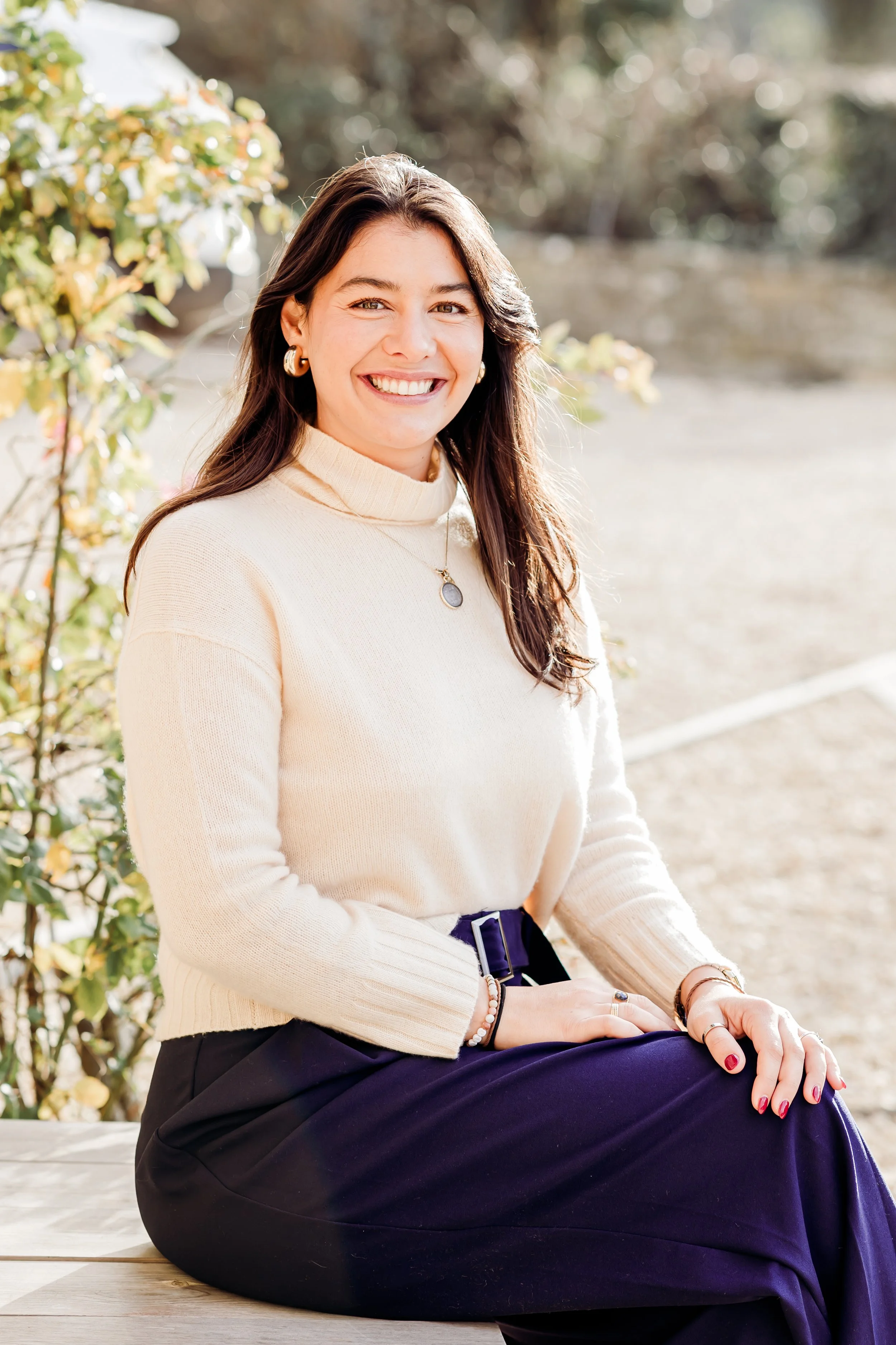 A woman with dark brown hair is smiling while sitting outdoors on a wooden surface, wearing a cream turtleneck sweater and dark pants, with greenery and sunlight in the background.