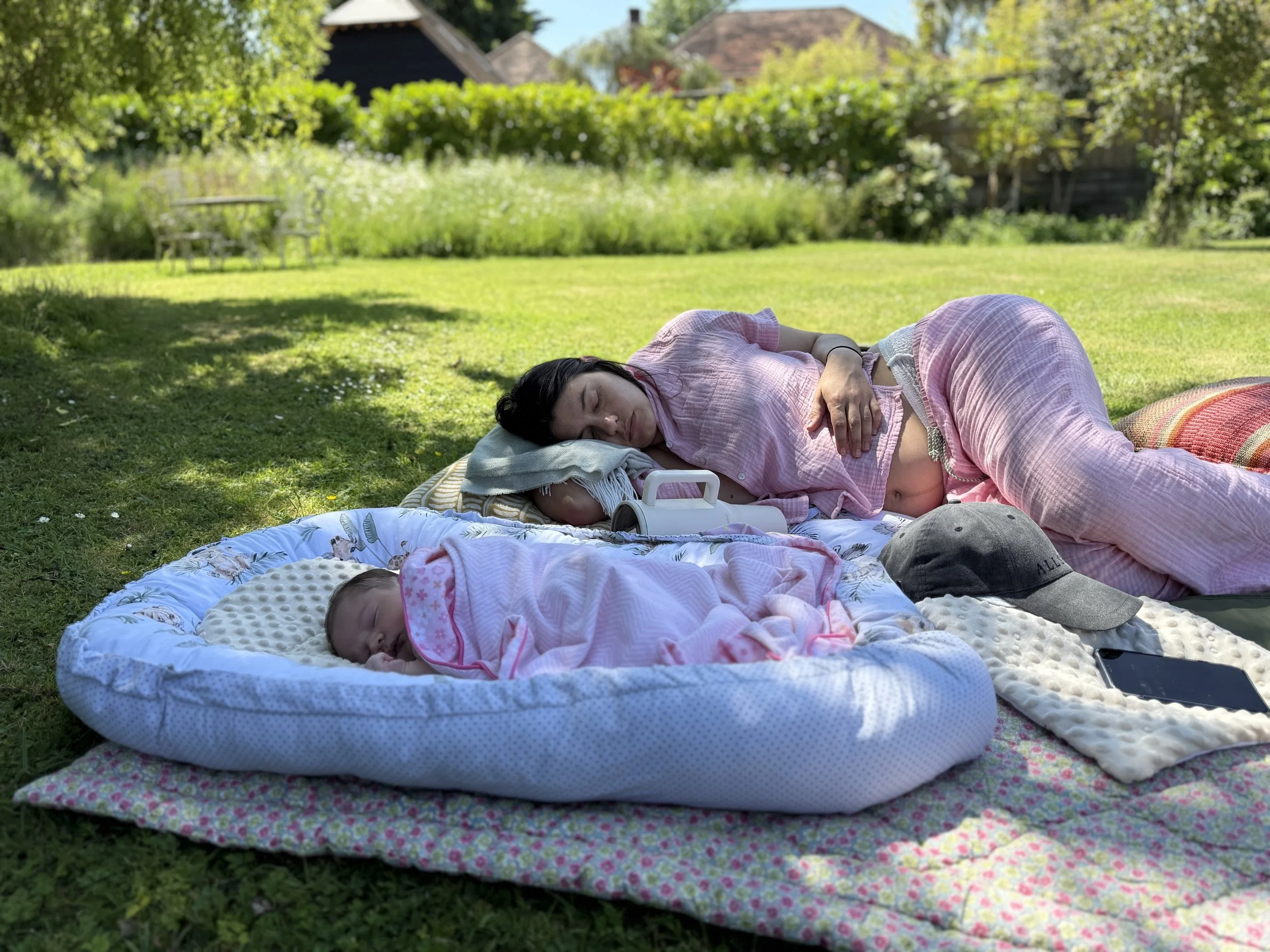 A woman and a baby sleeping outdoors on a grassy lawn with blankets under a tree, with houses and trees in the background.