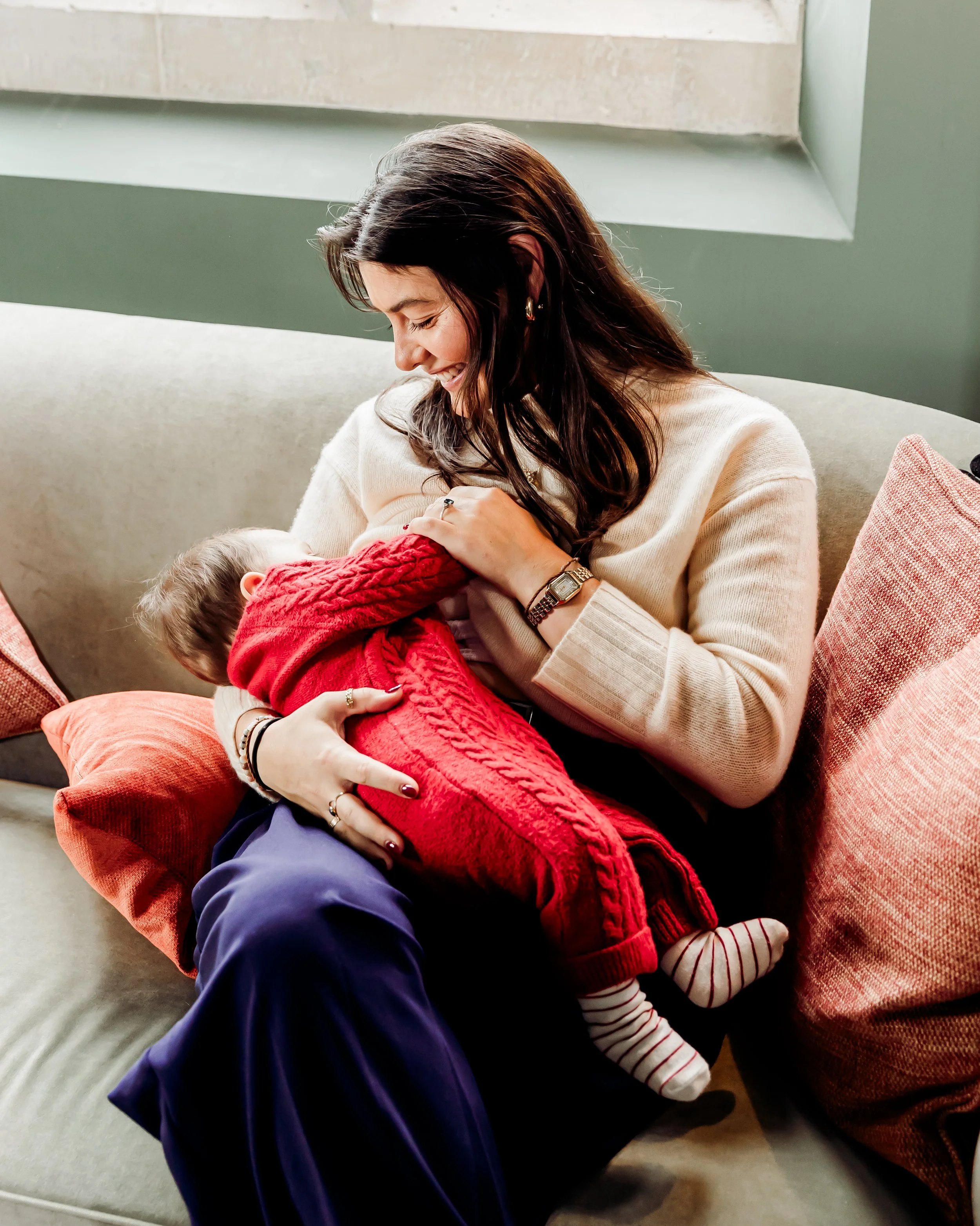 A woman with dark hair wearing a beige sweater and watch sitting on a sofa, smiling while cuddling a young child in a red sweater and striped pants.