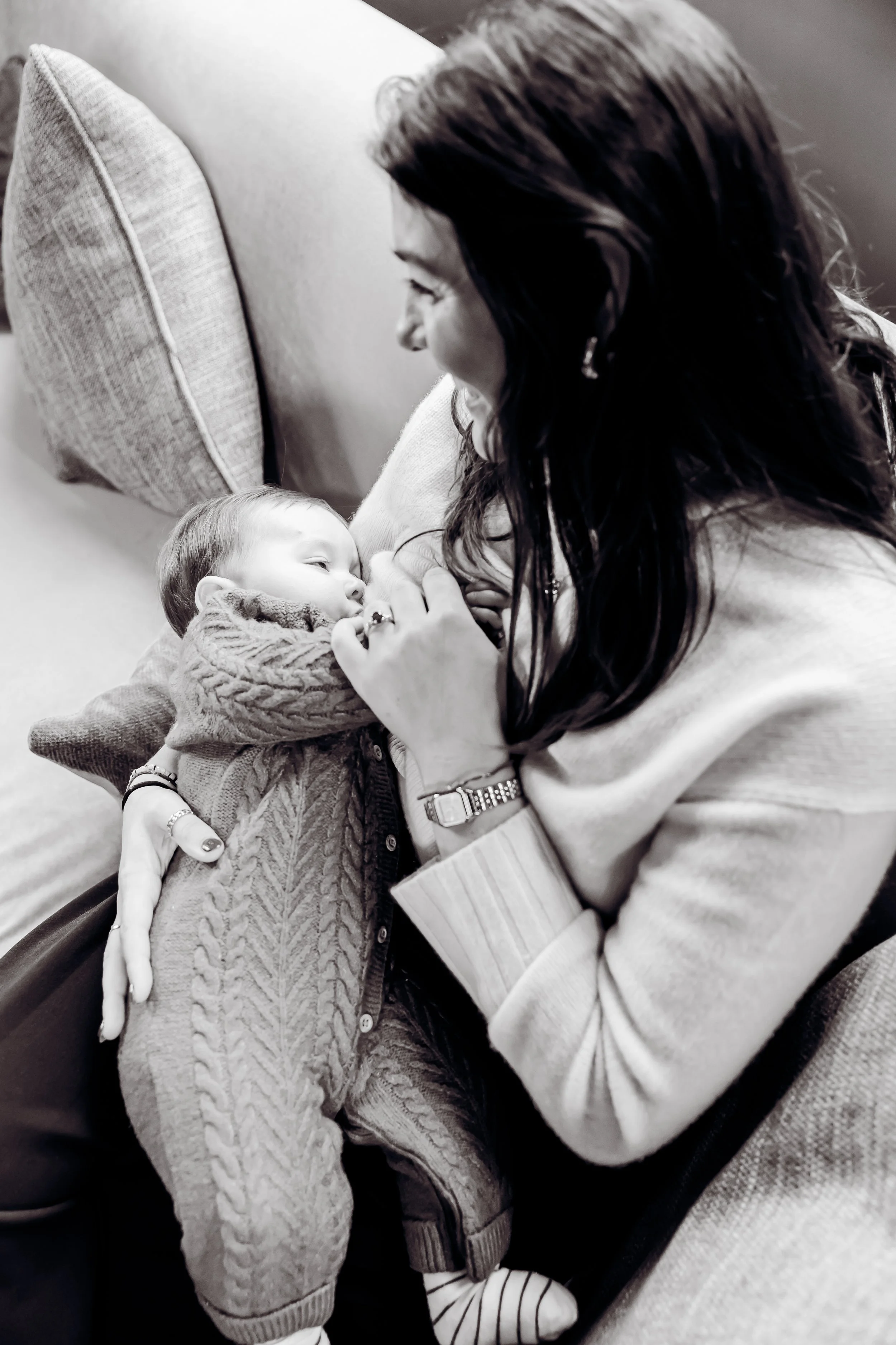A woman with dark hair and wearing a watch holds a young child with light hair, dressed in a cable-knit sweater, who is lying on her lap and gazing at her. They are sitting on a couch with pillows behind them.