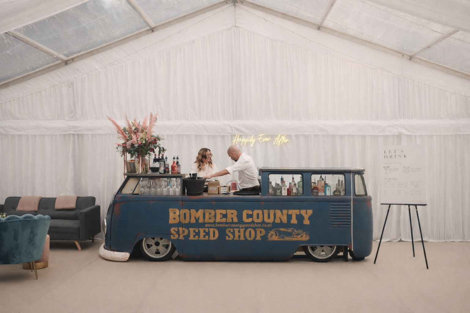 Luxury mobile bar styled beneath a light-filled marquee with draped fabric backdrop, curated florals, and relaxed lounge seating at a private event.