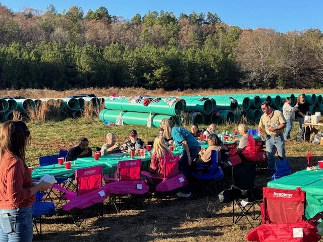 People gathered outdoors at a party with long tables and chairs, with large green pipes and wooded hills in the background.