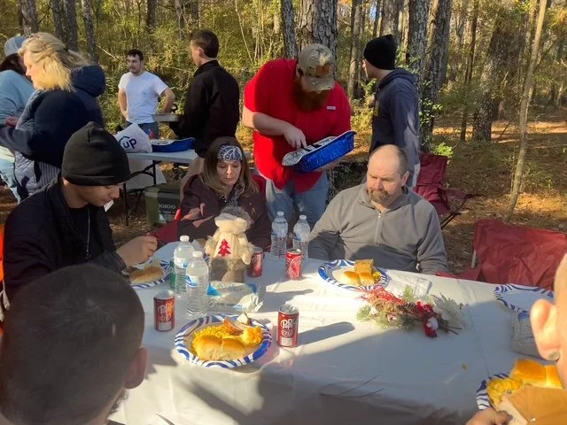 People gathered outdoors for a meal, with a table set with hot dogs, drinks, and holiday decorations.
