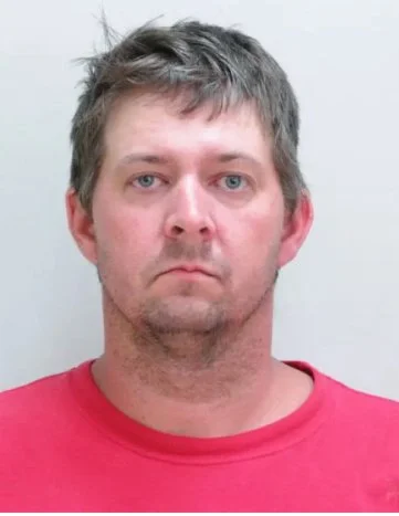 A man with short, light brown hair and a slight beard, wearing a red shirt, facing the camera with a neutral expression.