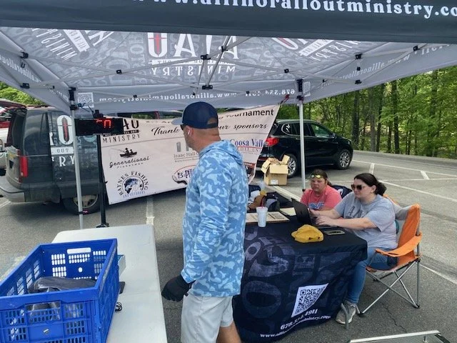 A group of people under a tent at an outdoor event, with a man in a blue patterned hoodie and baseball cap standing near a table. Behind him are two women sitting at a table with laptops, and a black car parked in the background. The tent has various