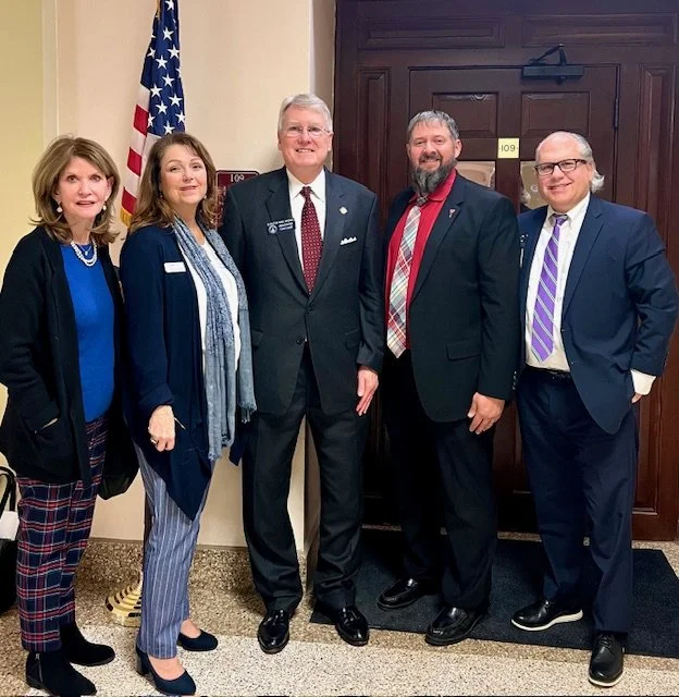Five professionally dressed people standing together indoors near an American flag, smiling for the camera.