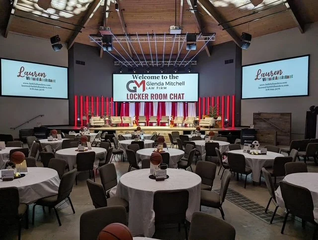 Event space set up with round tables and chairs, a stage with a large screen displaying 'Welcome to the Glenda Mitchell Law Firm Locker Room Chat,' and two smaller screens showing similar information. The stage has chairs and potted plants, and the v
