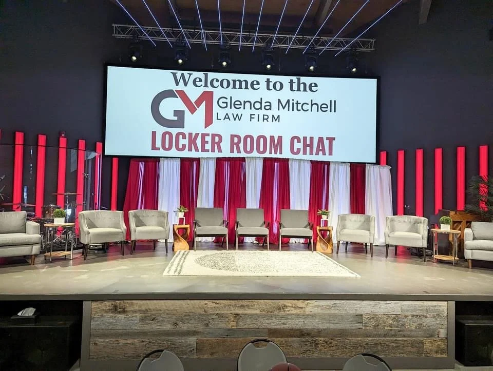 Stage setup for a panel discussion with nine chairs, side tables with plants, and a large screen displaying the logo and text for a law firm event titled 'Locker Room Chat'.