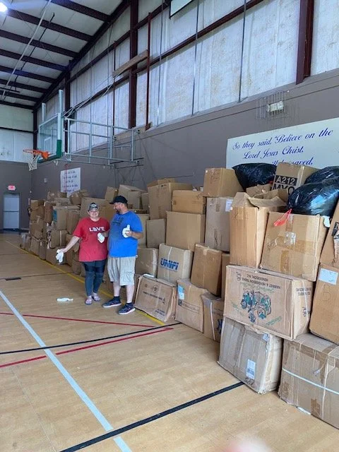 Two people standing in a gymnasium filled with stacks of cardboard boxes for a charity event.