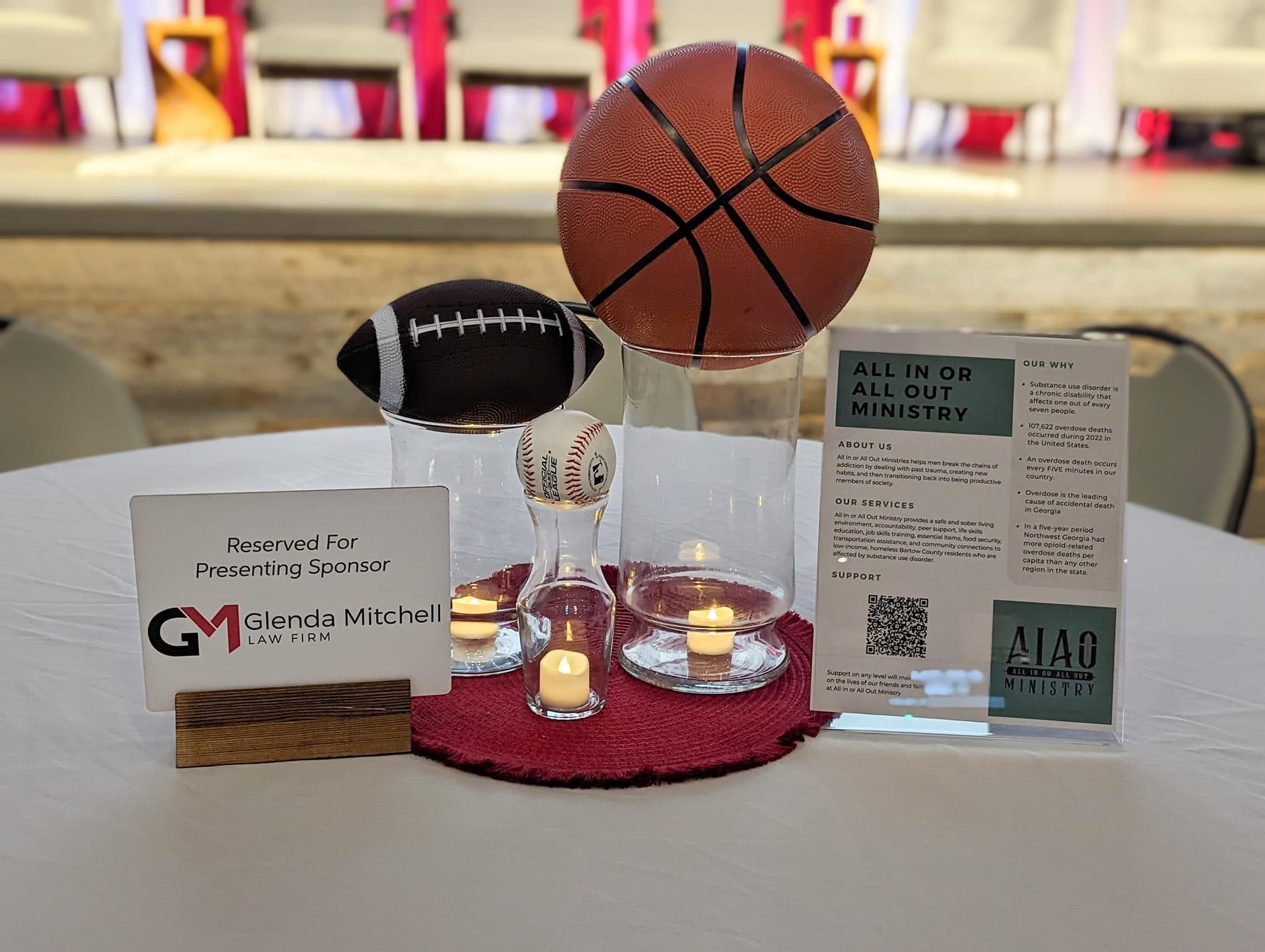 Table display with sports-themed decorations including a basketball, football, and baseball, each held by small glass stands. A small candle is lit in front of the display. A sign indicates it is reserved for Glenda Mitchell Law Firm. A flyer about a