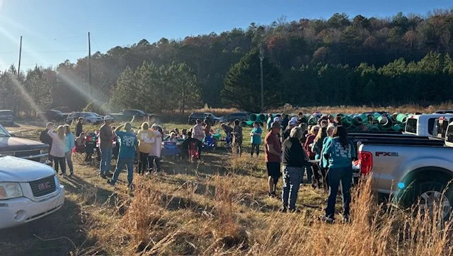 Group of people gathered outdoors in a grassy field, with cars parked nearby and trees in the background, some pipes or long objects on the ground.