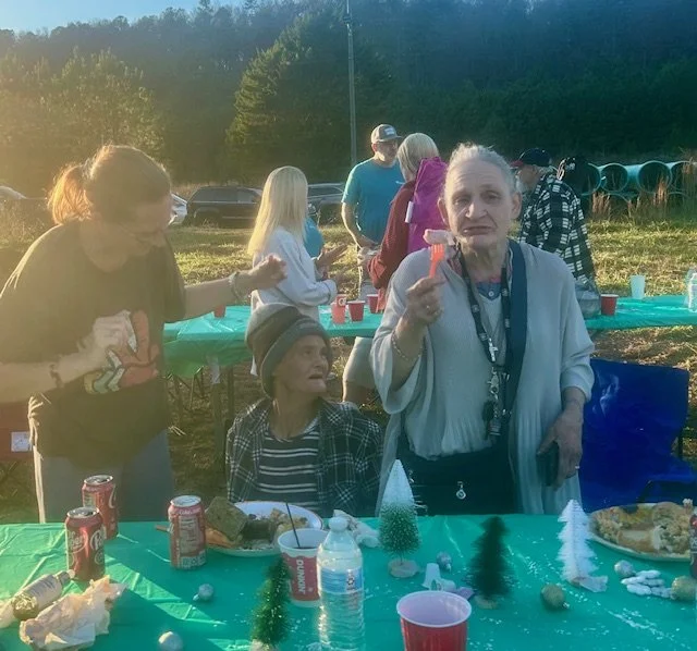 Group of people celebrating outdoors with a table of snacks and drinks, some sitting and some standing, during daytime.