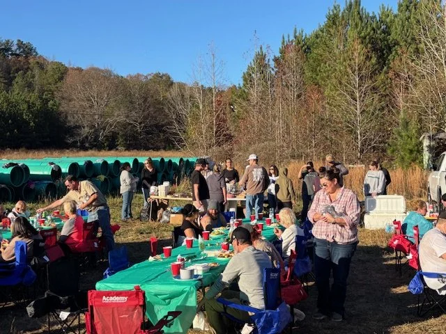 People gathered outdoors around tables with food and drinks, in a natural setting with trees and large green pipes in the background.