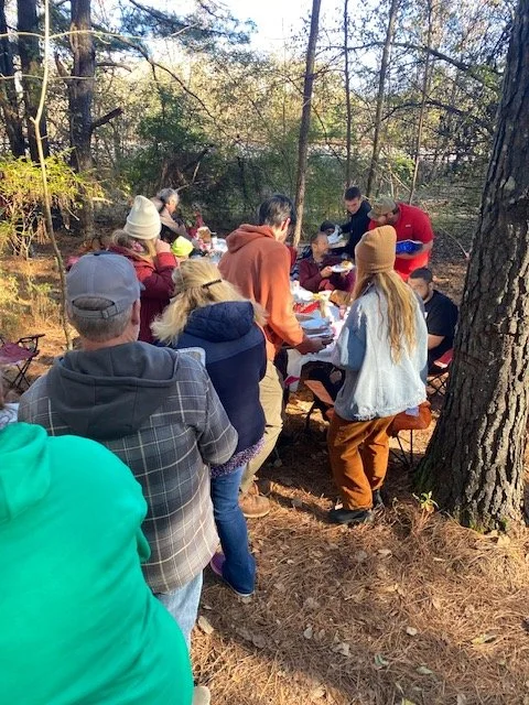 Group of people gathered around a table outdoors in a forest setting, enjoying a meal together.
