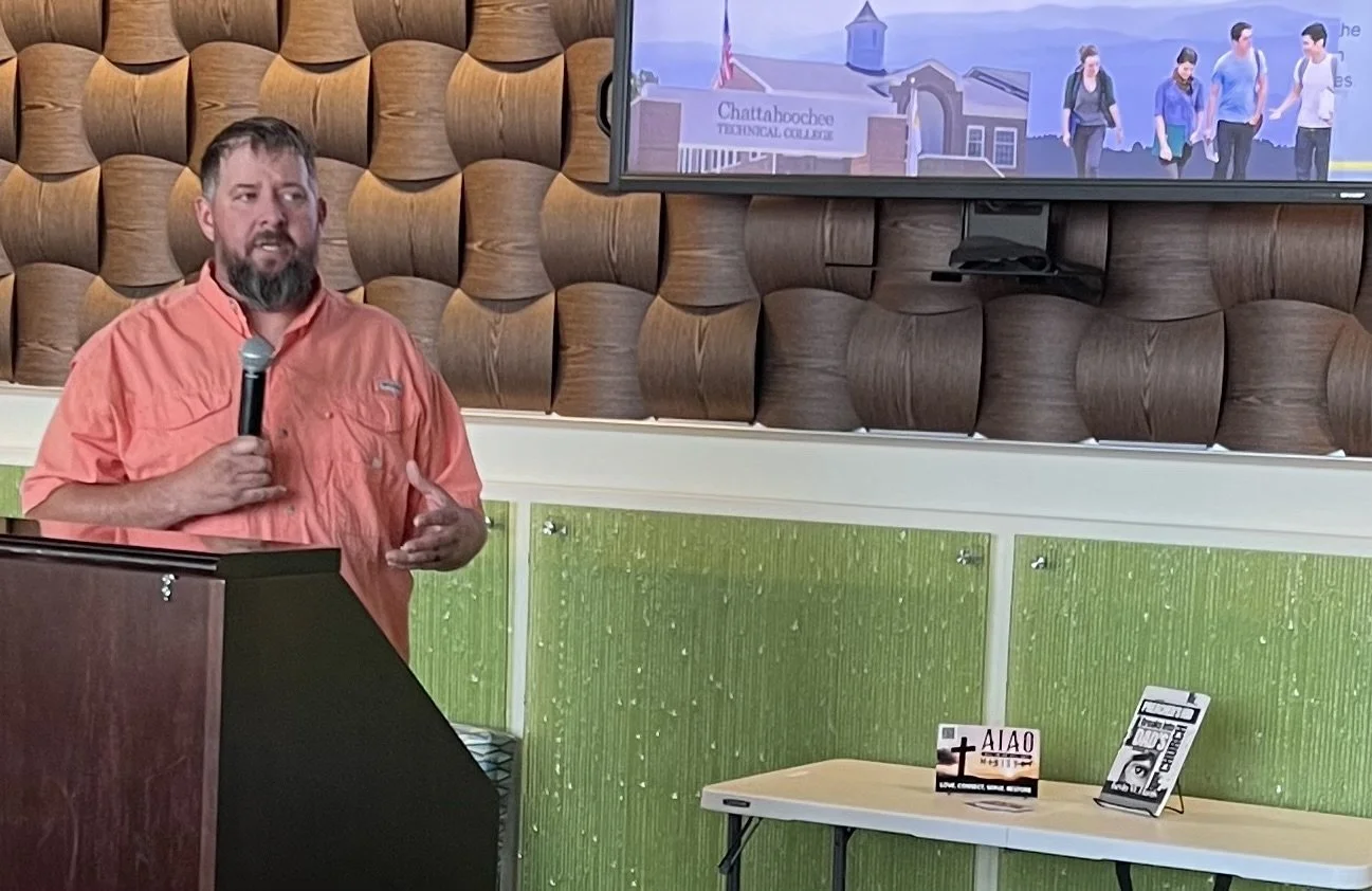A man speaking into a microphone at a presentation or lecture, with a television screen above him displaying a campus scene and a table with Christian informational materials.
