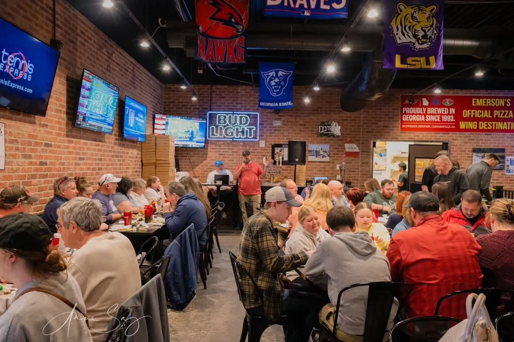A crowded restaurant with brick walls and multiple television screens, some displaying sports and advertisements, with a DJ or karaoke setup in the background. People are dining and socializing at tables.