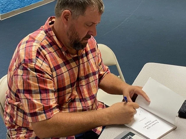 A man with a beard wearing a red plaid shirt, sitting at a table, signing a book.