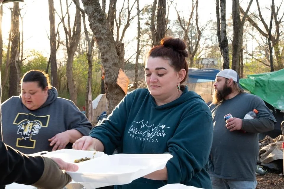 Group of three people outdoors, with trees and tents in the background. One woman in the center is serving food on a white foam plate, wearing a blue hoodie. Another woman on the left is wearing a gray hoodie with a logo, and a man on the right is ho