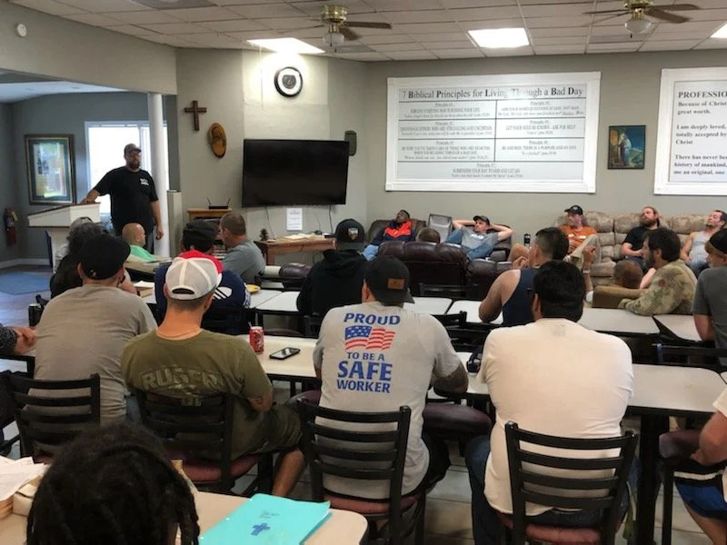 Group of men attending a presentation or seminar in a room with walls decorated with religious and biblical posters. A speaker stands at the front, and a large television screen is mounted on the wall.