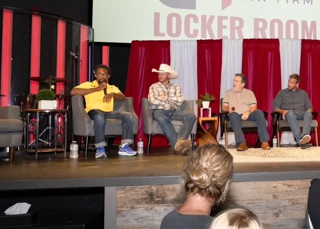 Four men sitting on chairs on a stage during a panel discussion, with one man speaking into a microphone. Behind them is a backdrop with red and white drapes and a sign that says 'LOCKER ROOM.' A woman with blonde hair tied in a bun is seated in the 