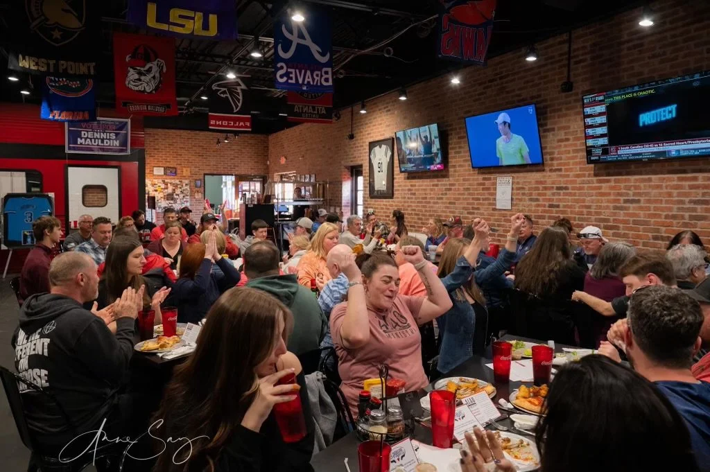 Crowd of people sitting at tables in a sports bar watching television screens, some celebrating with raised arms and clapping.
