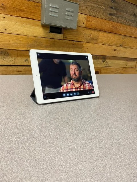 A tablet on a table displays a man with a beard and a plaid shirt, with a wooden wall and an electrical box in the background.