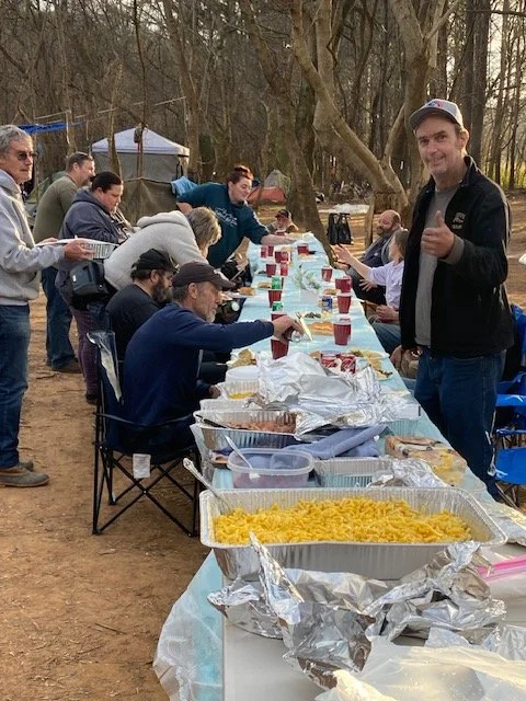 People gathered outdoors around a long buffet table filled with food and drinks, enjoying a meal together.