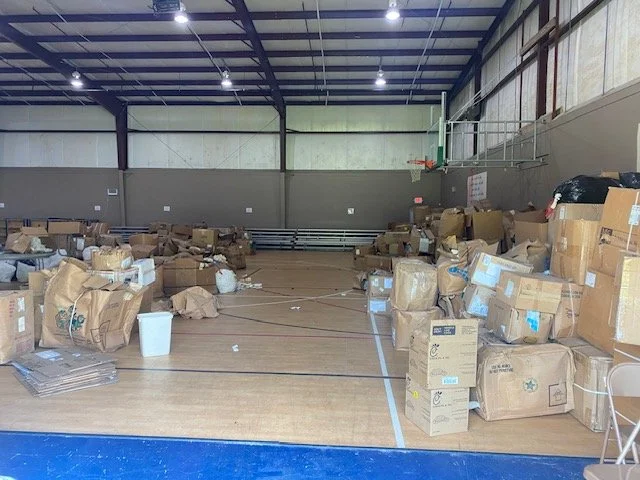 An indoor gymnasium filled with numerous cardboard boxes and packages scattered across the wooden floor, with a basketball hoop and backboard mounted on the wall.