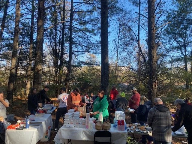 People gathering at an outdoor event in a wooded area, serving food from tables lined up in front of trees.