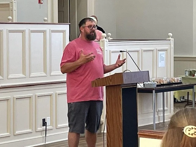 A man with glasses and a beard, wearing a pink T-shirt and shorts, speaking at a podium in a room with white paneling and a table with food.
