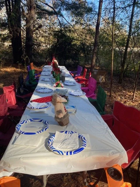 An outdoor table set for a celebration in a forested area with colorful chairs, decorated with napkins and a plush toy at the center.