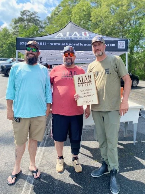 Three men standing outdoors in front of a tent, holding a plaque that reads 'AIAO Ministry 1st Annual Fishing Tournament 1st Place.' The man in the middle is wearing a red shirt, black shorts, and sunglasses. The man on the left is wearing a light bl